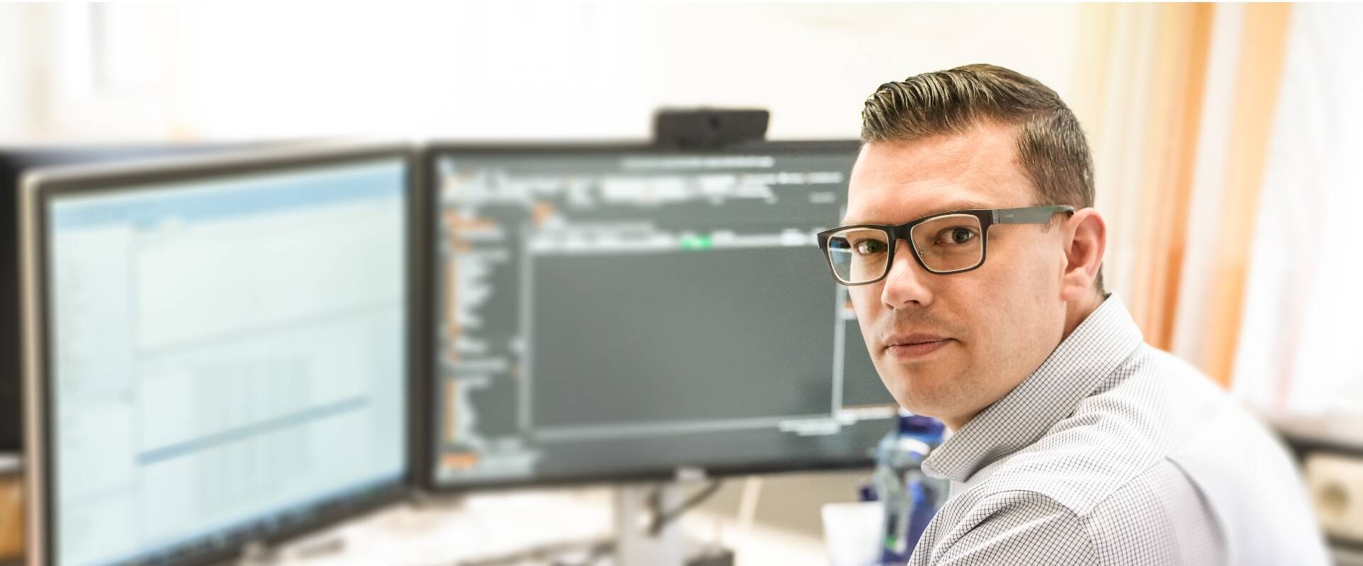 Freundlicher Mitarbeiter mit Brille sitzt im Büro vor 2 Bildschirmen